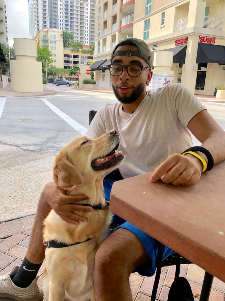 Man wearing glasses and cap sitting at a table with a yellow Labrador dog on an urban street